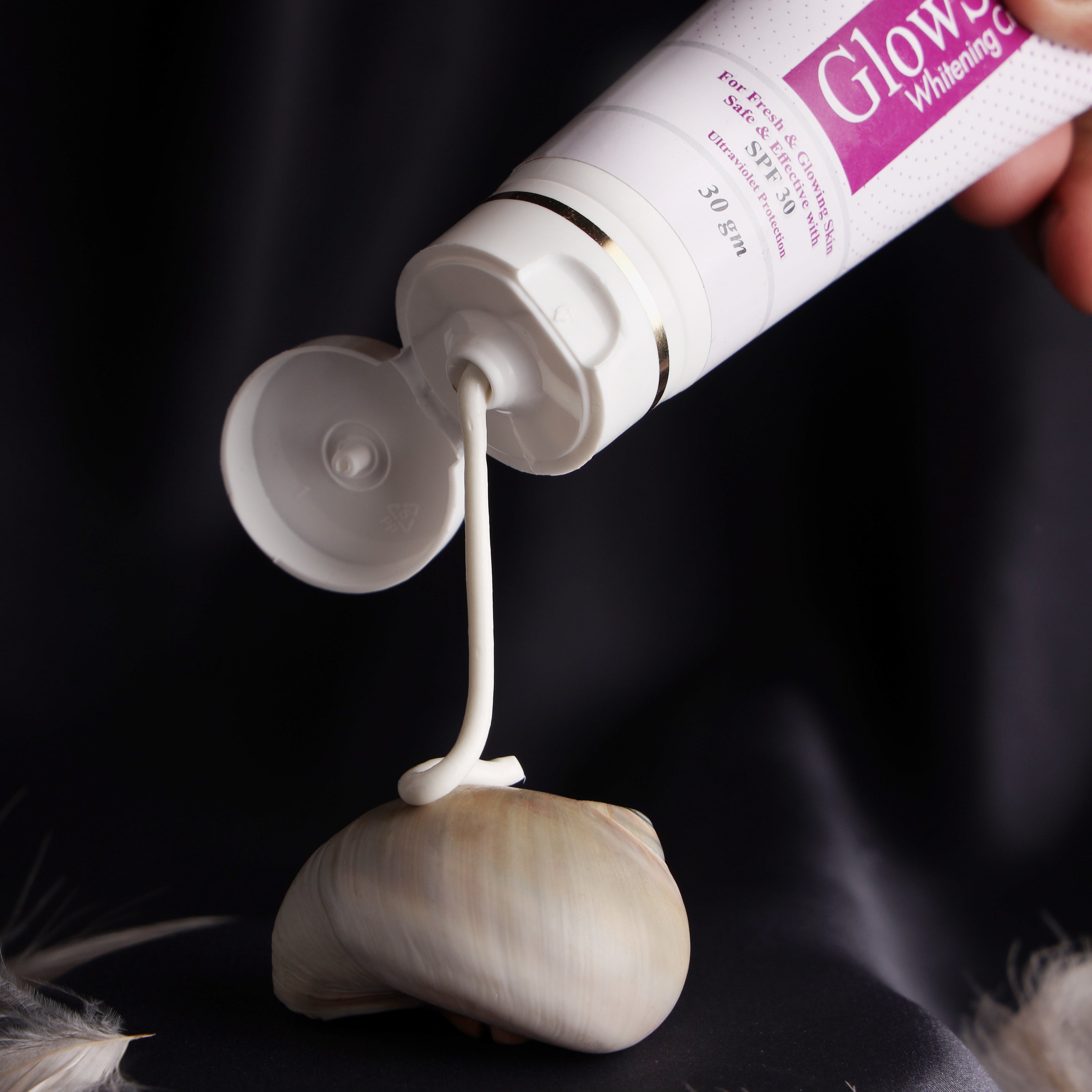 Skin care cream tube dispensing lotion onto a seashell, dark background