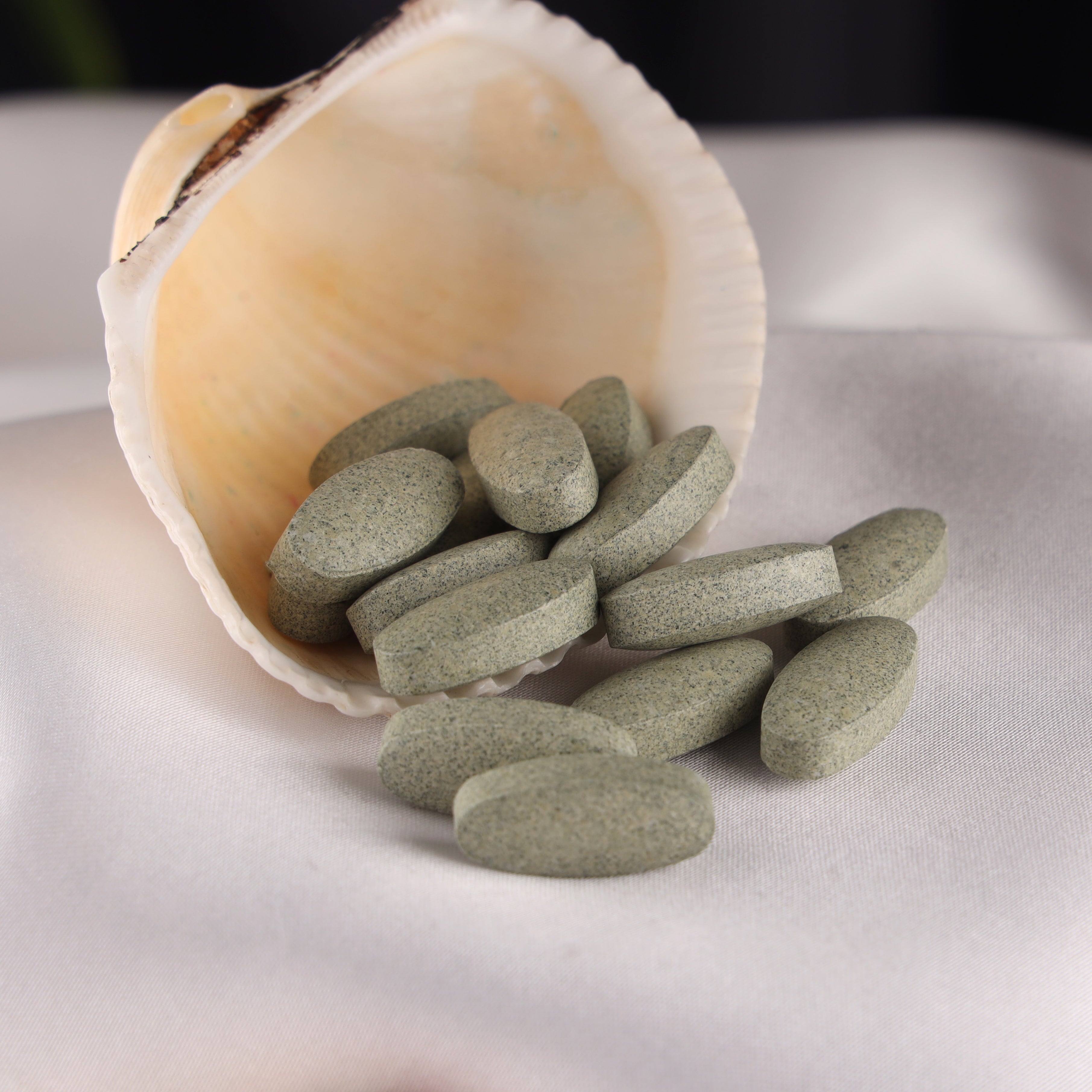 Green supplement tablets spilling from a seashell onto a soft white fabric surface
